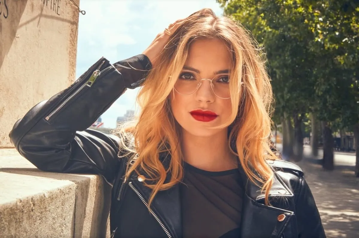 Young woman with blonde wavy hair, red lipstick, clear round glasses, and a black leather jacket leaning against a stone wall outdoors.