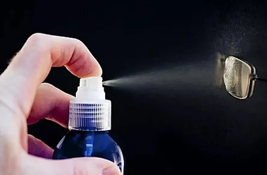 Hand spraying cleaning solution onto one lens of eyeglasses against a black background.
