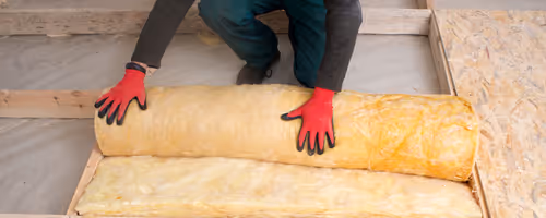 Person wearing red gloves rolling out yellow fiberglass insulation between wooden frame studs on a floor.