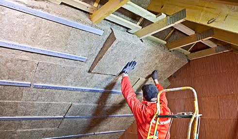 Worker in red jacket installing brown insulation panels on a roof attic ceiling.