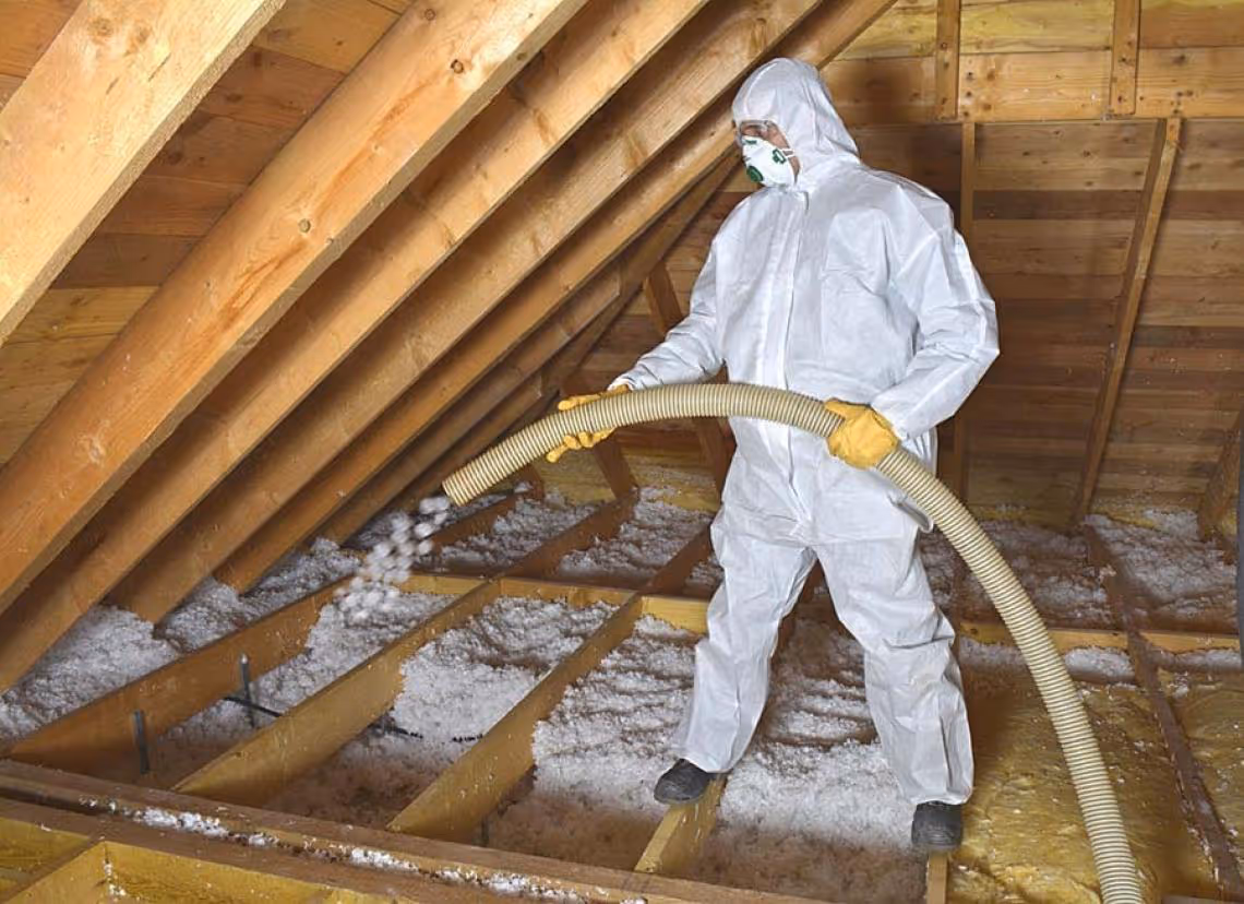 Person in white protective suit and mask installing loose-fill insulation in an attic using a hose.