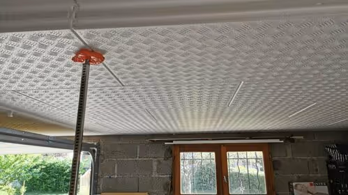 Close-up of a textured white ceiling with a red adjustable support jack holding it up in a garage with a window and concrete block walls.