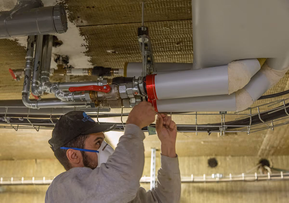 Worker wearing a mask and cap installing or repairing insulated pipes with red valve handles on a ceiling.