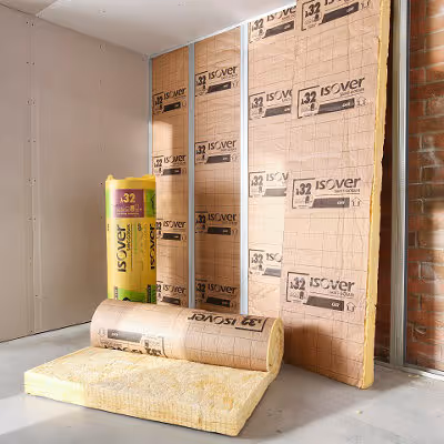 Yellow Isover insulation rolls partially installed between wooden wall studs in an unfinished room.