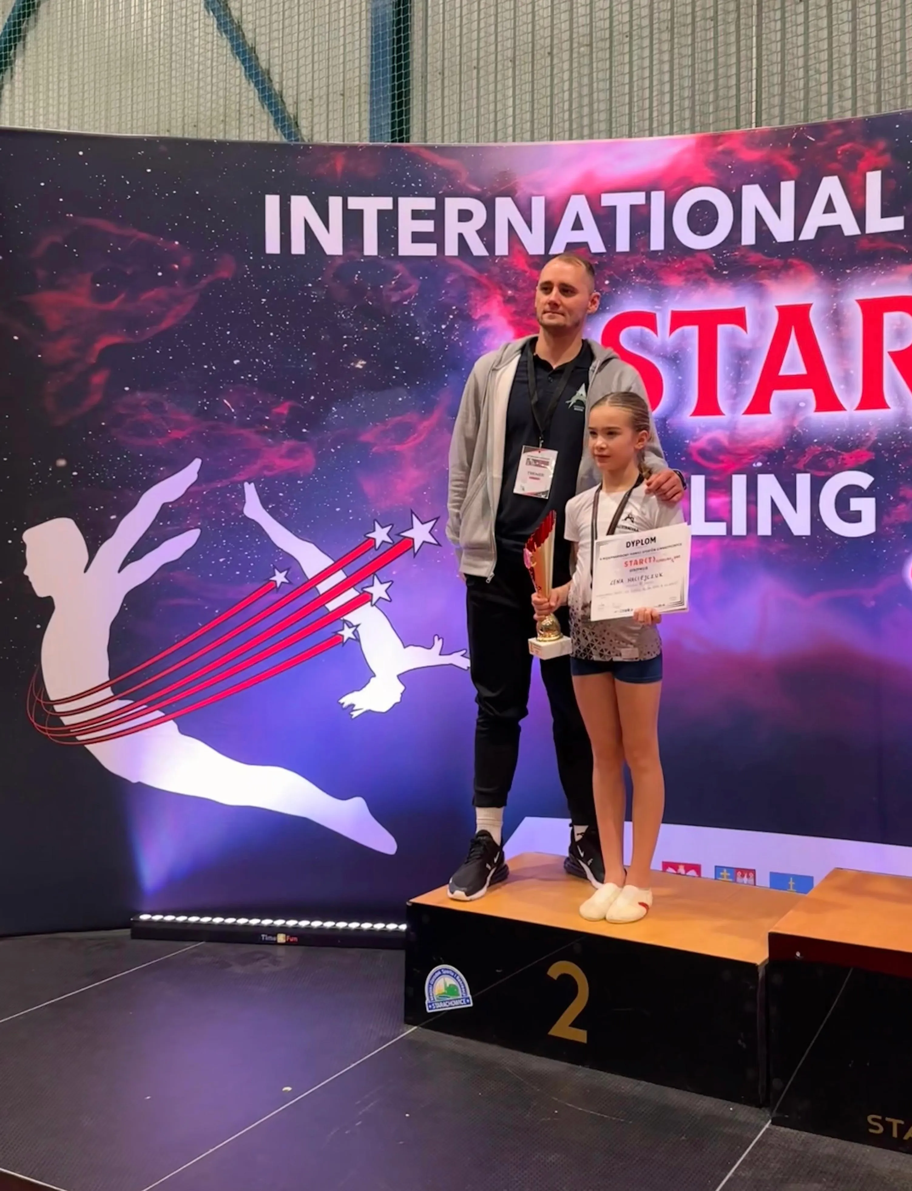 Młoda gimnastyczka na podium wraz ze swoim trenerem.