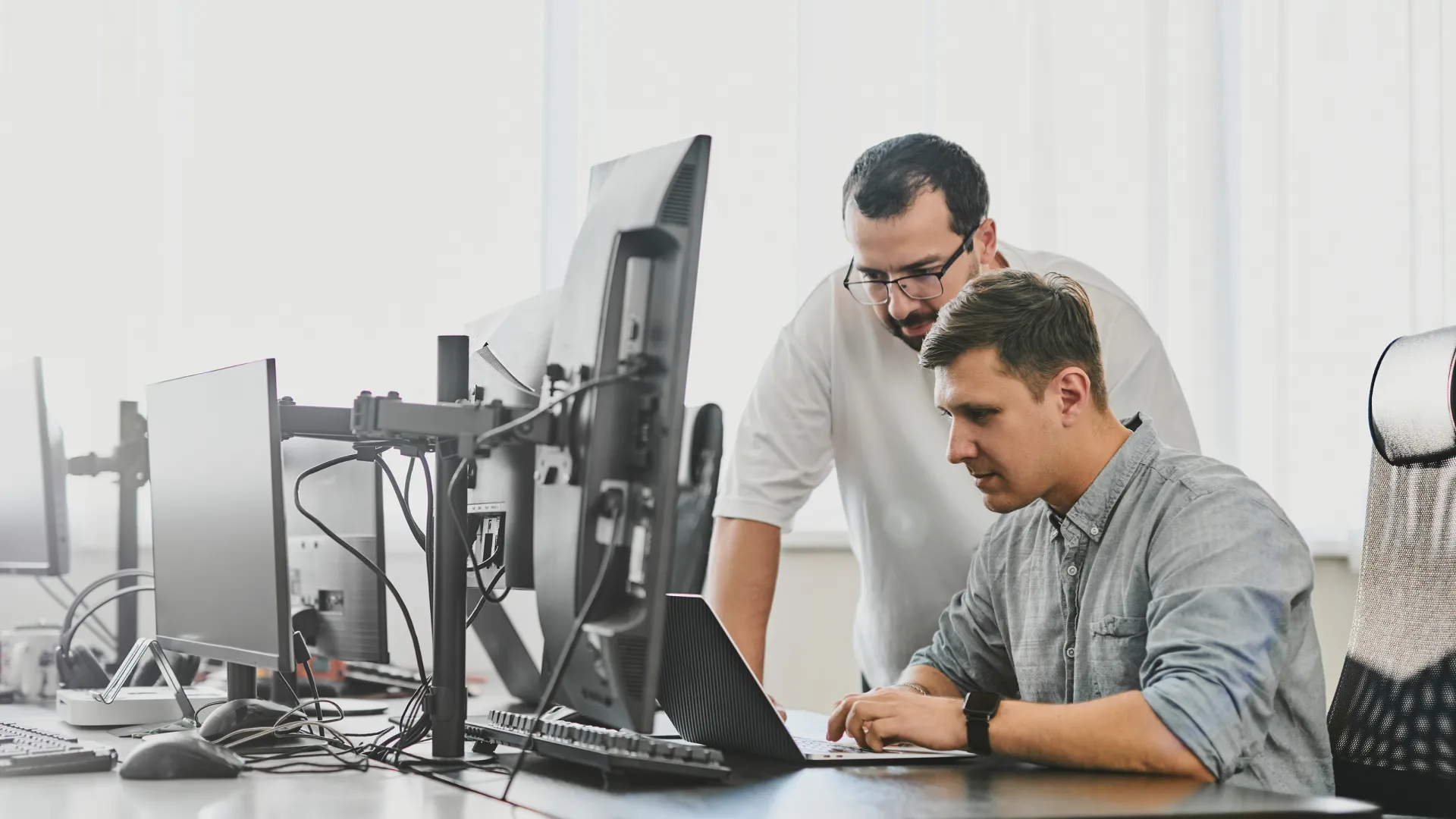 Two men working together at a desk with a laptop and multiple monitors in an office.
