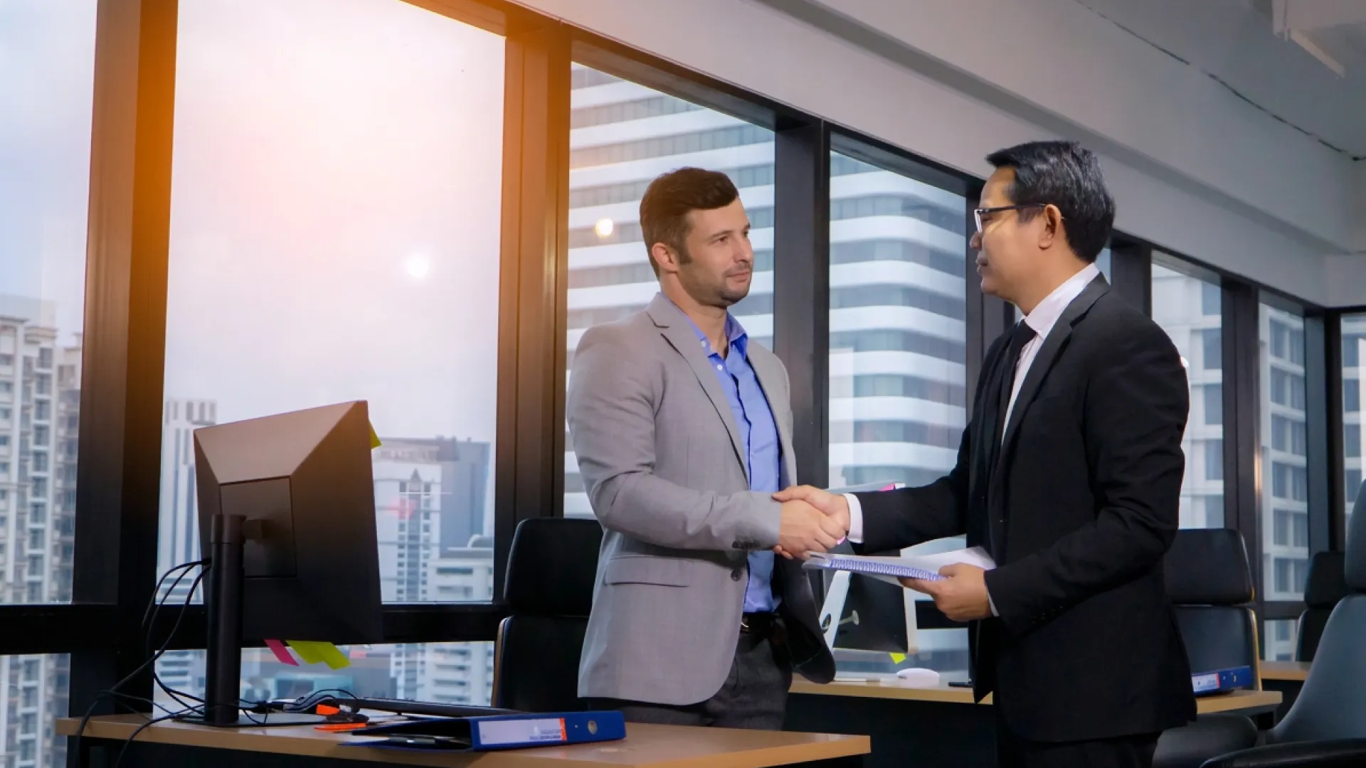 Two businessmen shaking hands in a modern office with city buildings visible through large windows.