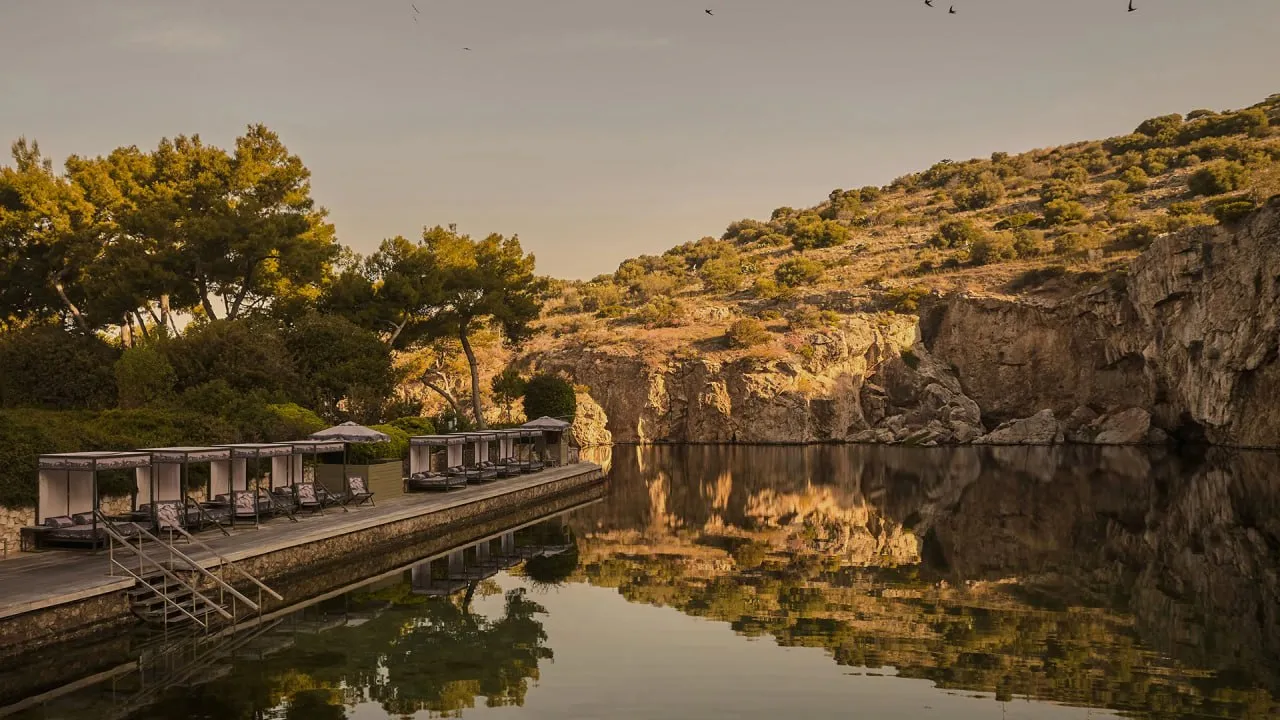 Wooden poolside deck with lounge beds next to calm water reflecting rocky hillside and trees.