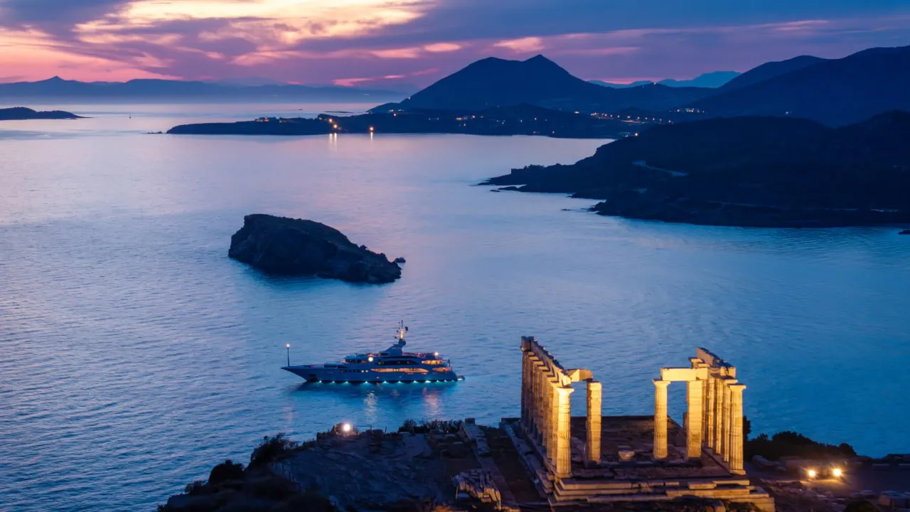 Illuminated ancient Greek temple ruins by a bay with a yacht on calm water at sunset.