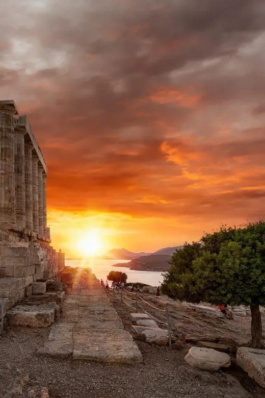 Sunset over ancient stone ruins with a tree and distant mountains by the sea.