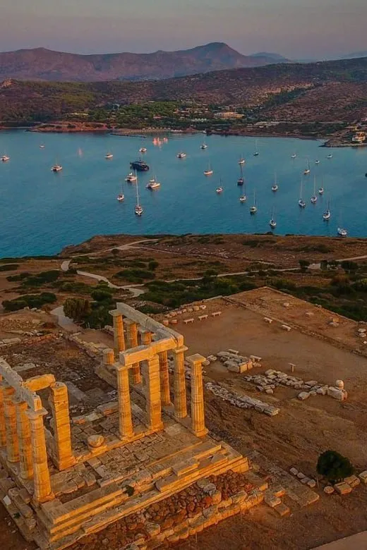 Ancient Greek temple ruins near the coast with boats anchored in blue water and hills in the background.