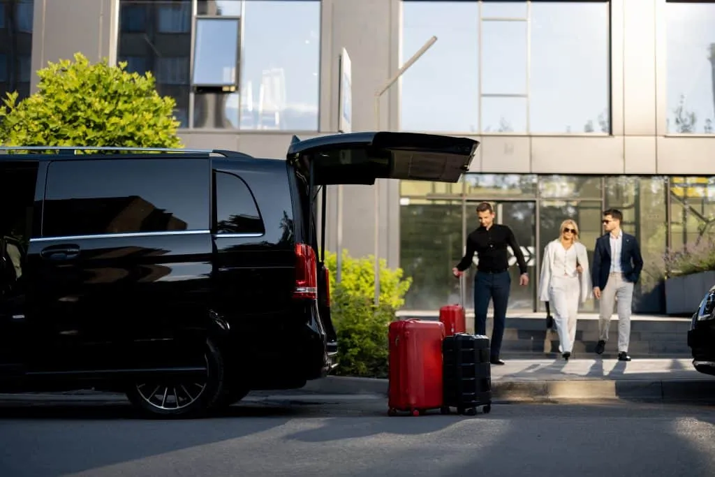 Three well-dressed people walking towards a black van with luggage at its open trunk.