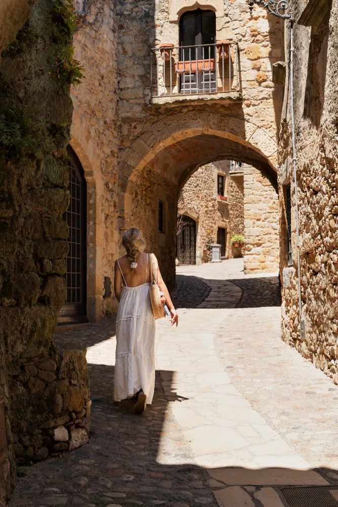 Woman in a white dress walking through a sunlit stone alley with arches in an old village.