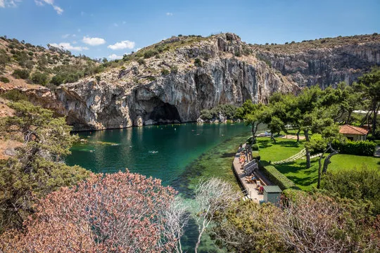 Scenic lake surrounded by cliffs and trees with a cave entrance visible in the rocky hillside.