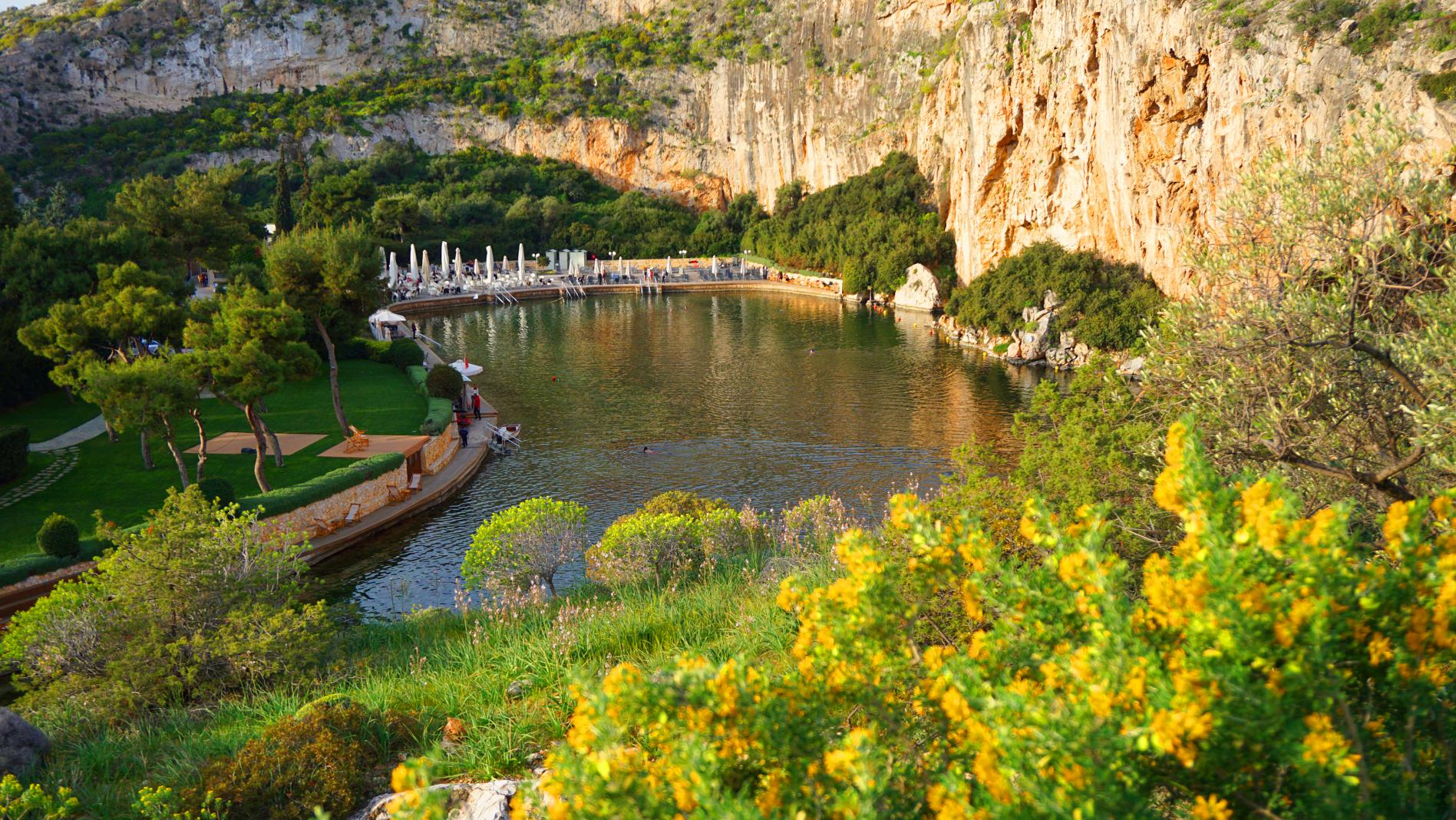 Lake surrounded by rocky cliffs, green trees, yellow flowers, and a dock with umbrellas and lounge chairs.