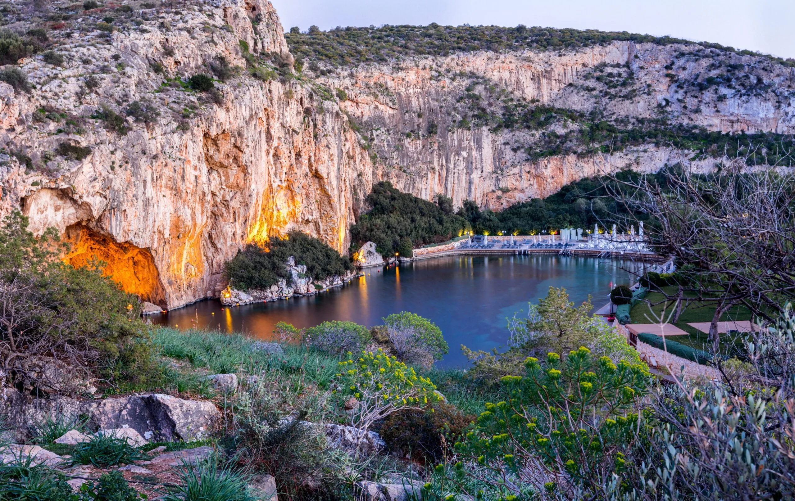 Lake surrounded by rocky cliffs and lush greenery with cave entrances illuminated by warm lights at dusk.
