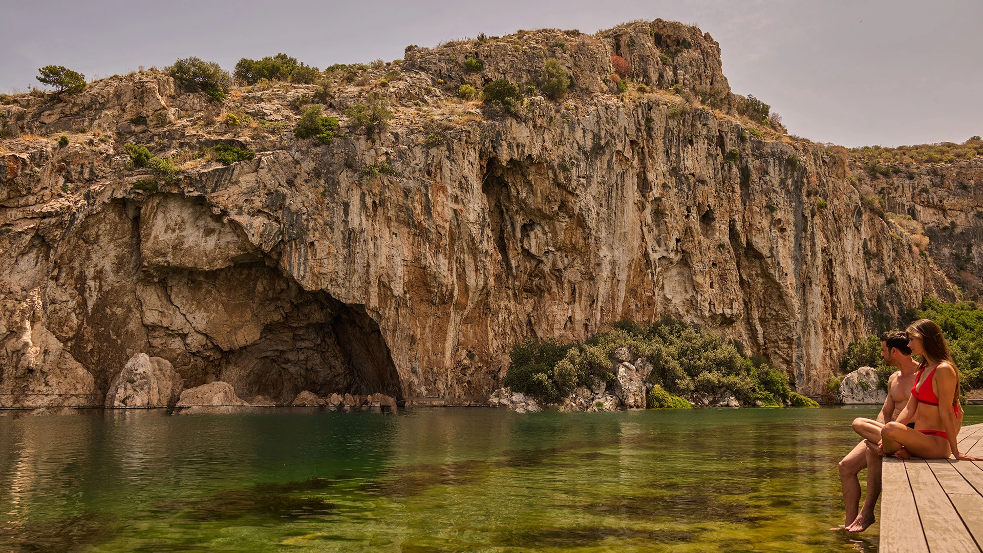 Couple in swimwear sitting on a wooden dock by clear water with a rocky cliff and cave in the background.