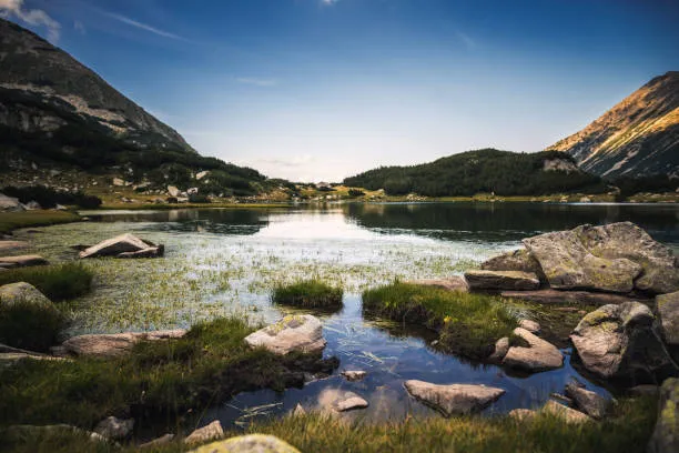 Mountain lake with clear water, surrounded by grassy rocks under a blue sky.