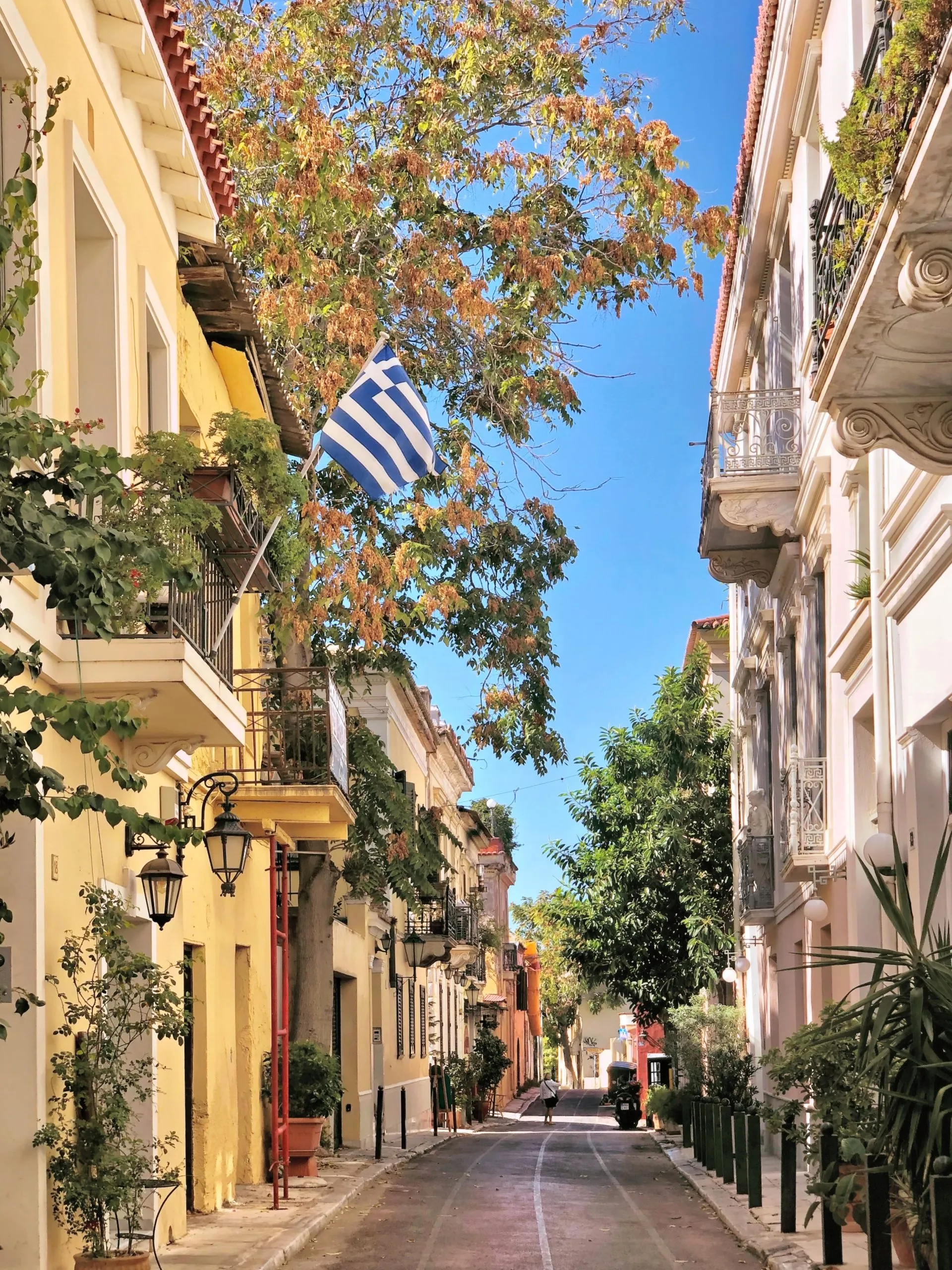 Narrow street with yellow and white buildings, a Greek flag, trees, and a person walking in the distance.
