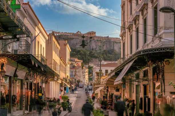 Busy pedestrian street with shops and historic buildings leading to a rocky hill with ancient ruins under a blue sky.