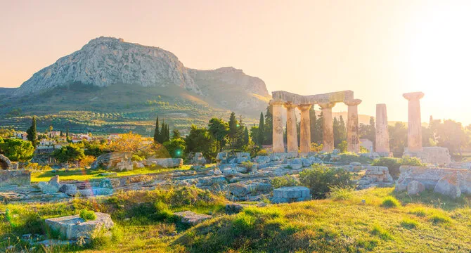 Ancient ruins with stone columns and scattered rocks under a mountain at sunset.
