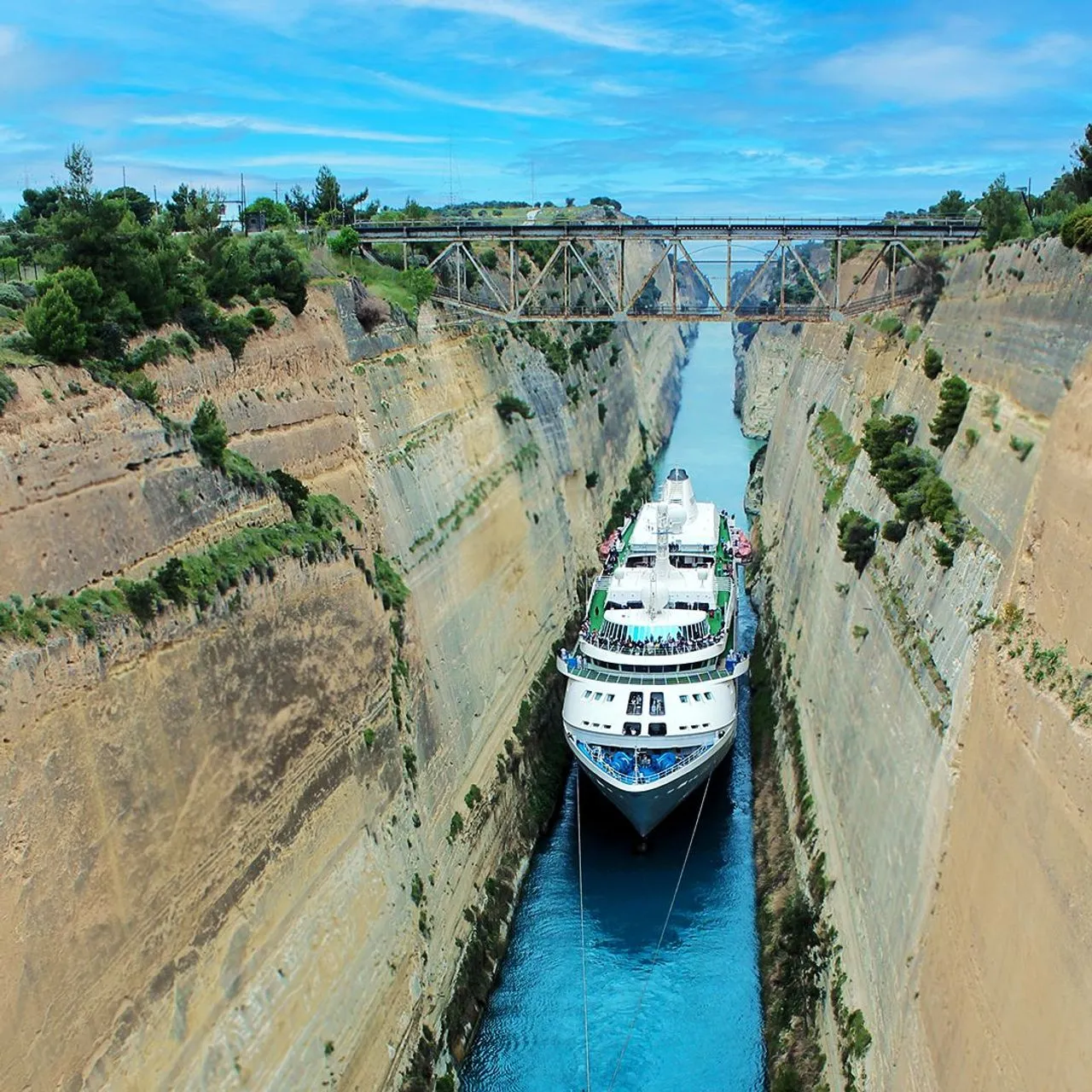 Large white cruise ship navigating narrow blue canal with steep rocky cliffs and a metal bridge overhead.