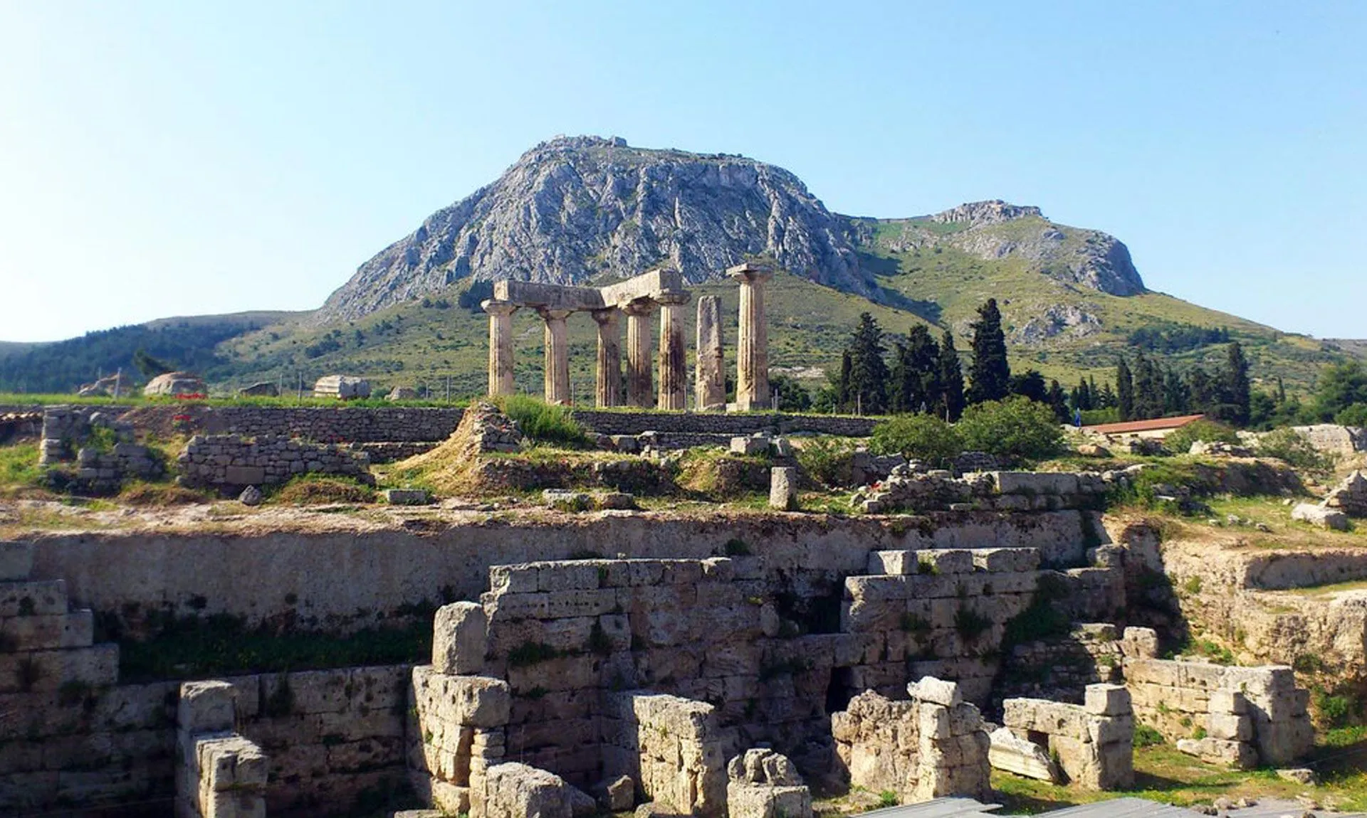 Ancient Greek ruins with stone columns set against a mountainous landscape under a clear blue sky.