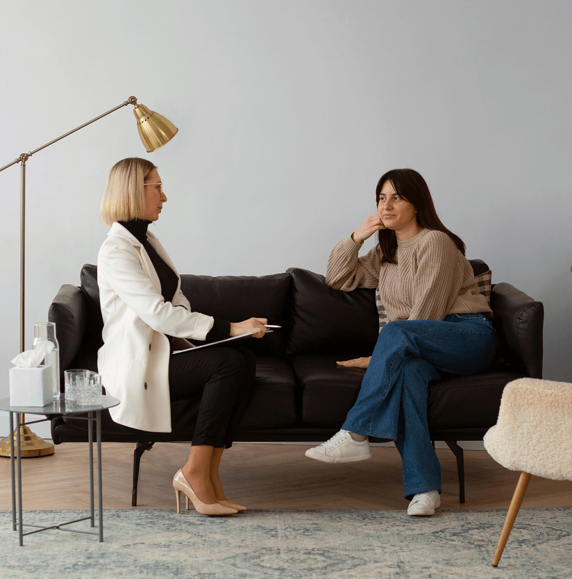 Therapist in white blazer talking with a woman in a brown sweater sitting on a black couch in a cozy room.