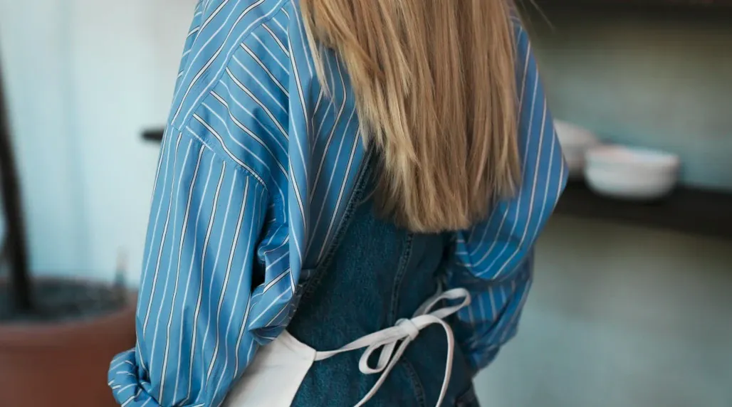Frau mit langem blonden Haar trägt blaue gestreifte Bluse und einen weißen Kittel mit Schleife am Rücken.