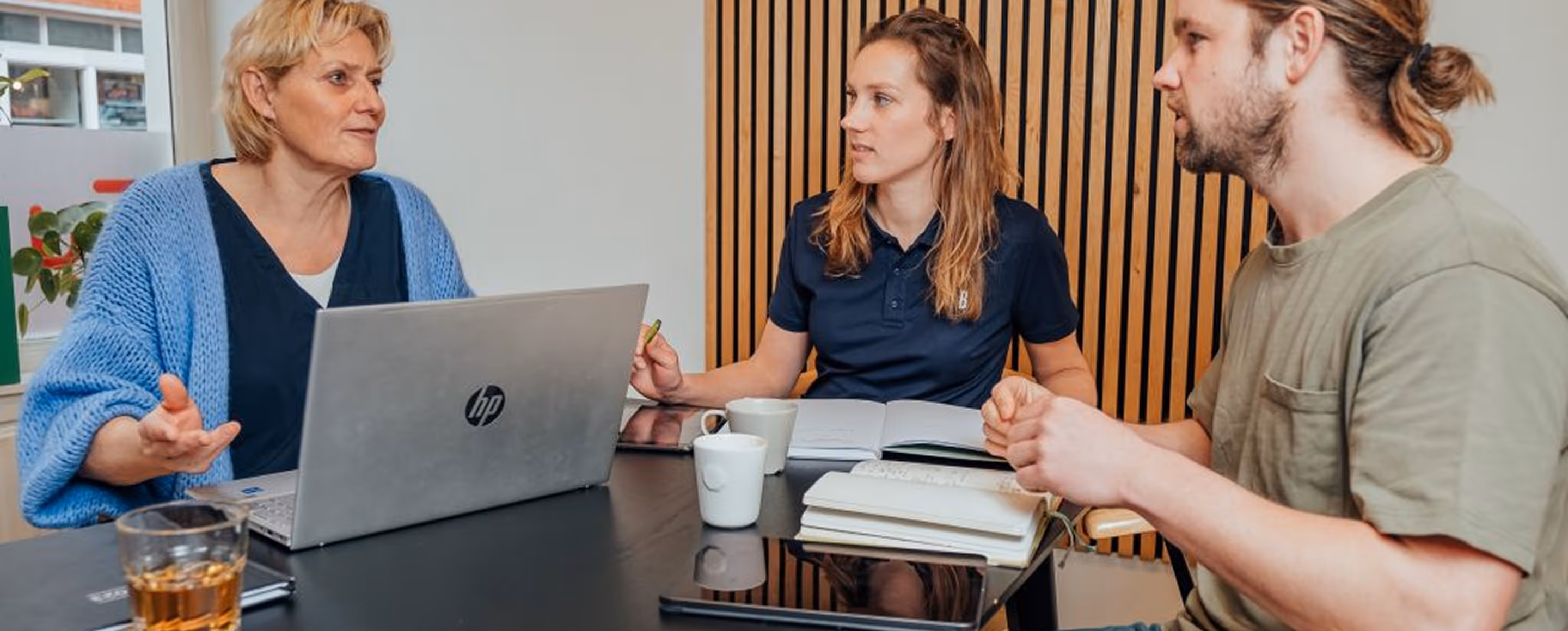 Drie personen in gesprek aan een tafel met een laptop, notitieboekjes en drinken.
