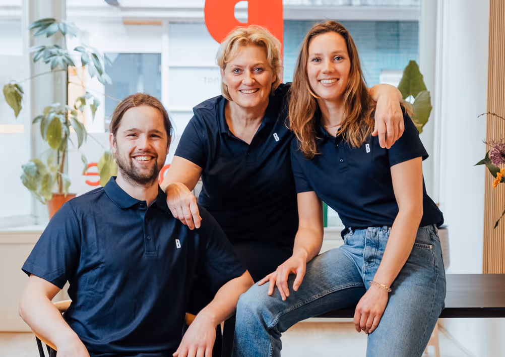 Drie personen in donkerblauwe polo's lachen samen in een lichte kamer met planten en een grote raamachtergrond.