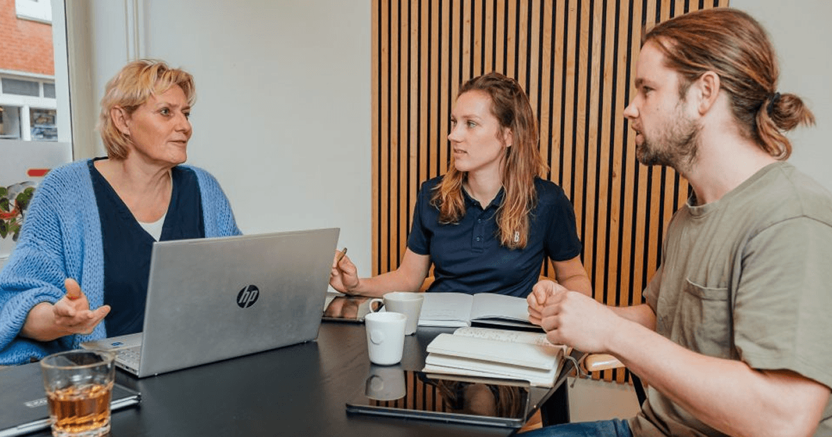 Drie volwassenen in gesprek aan tafel met laptop, notitieboekjes en kopjes koffie.