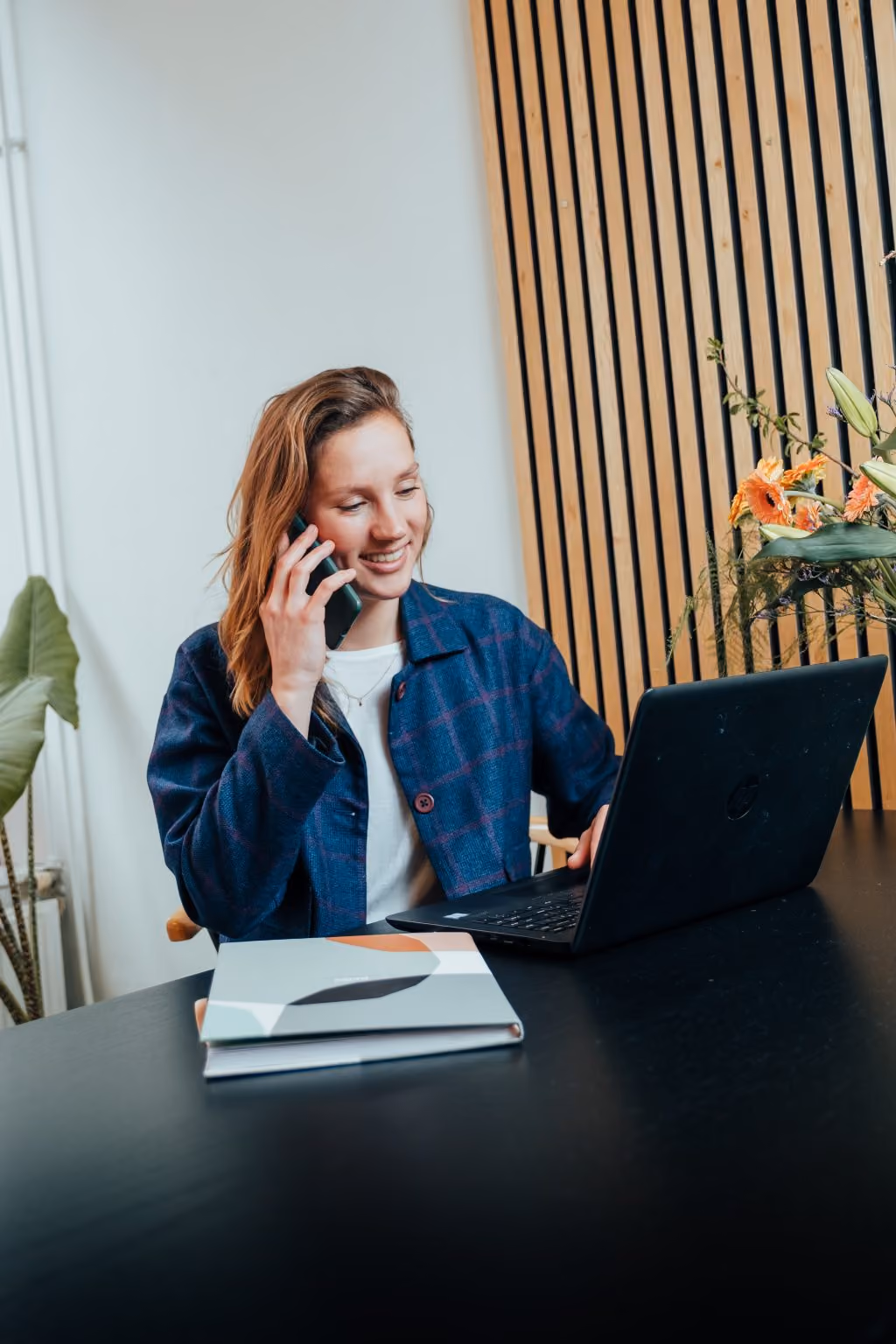 Vrouw aan tafel praat op telefoon terwijl ze op laptop werkt met bloemen op de achtergrond.