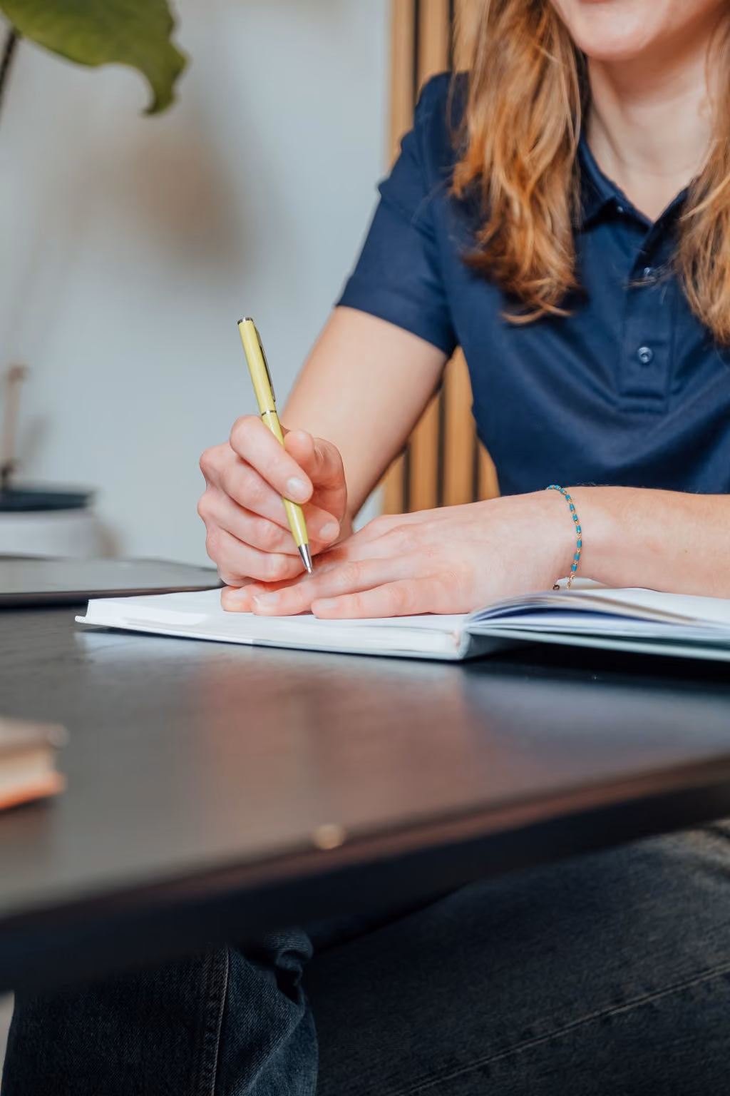 Vrouw in blauwe polo schrijft met gele pen in open notitieboek op tafel.