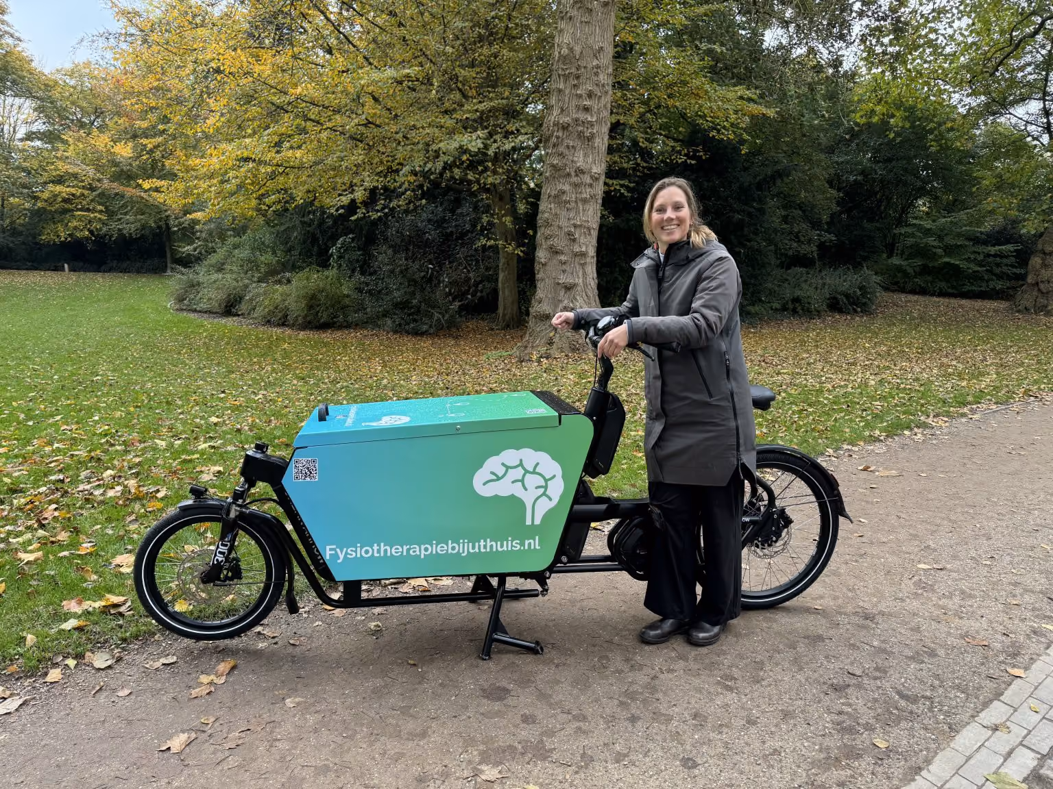 Vrouw in jas staat naast een bakfiets met het logo en tekst Fysiotherapiebijjuthuis.nl in een park.