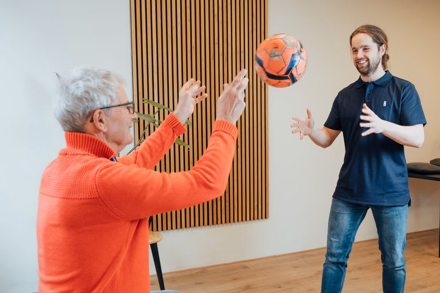 Ouder persoon in oranje trui en jongere man vangen een oranje bal in een lichte kamer.