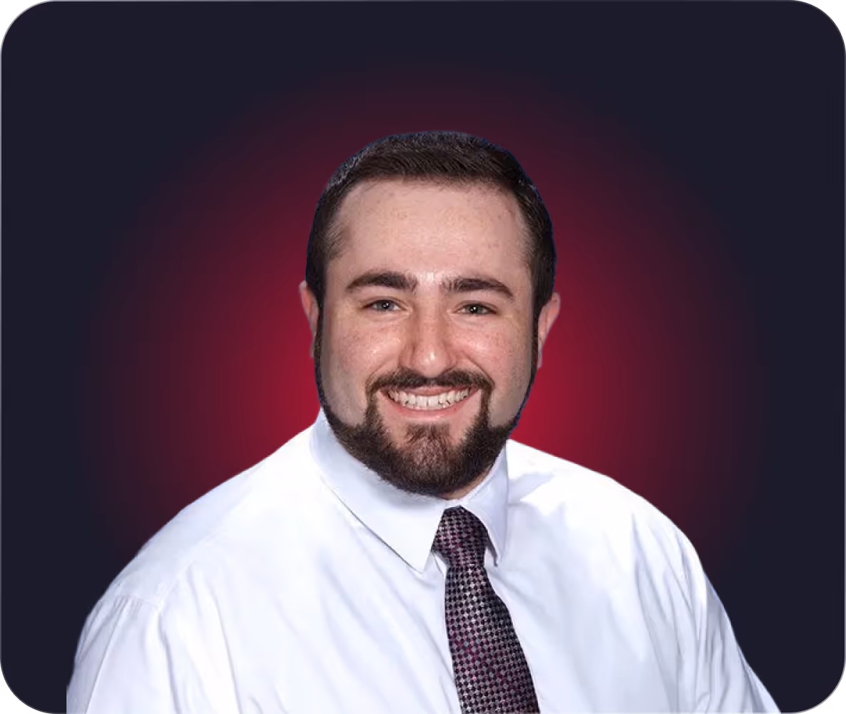 Smiling man with dark hair and beard dressed in white shirt and patterned tie against a dark background with a red gradient glow.