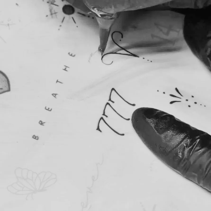 Close-up of a tattoo artist's gloved hand drawing a tattoo with the word 'BREATHE' and number '777' on skin.