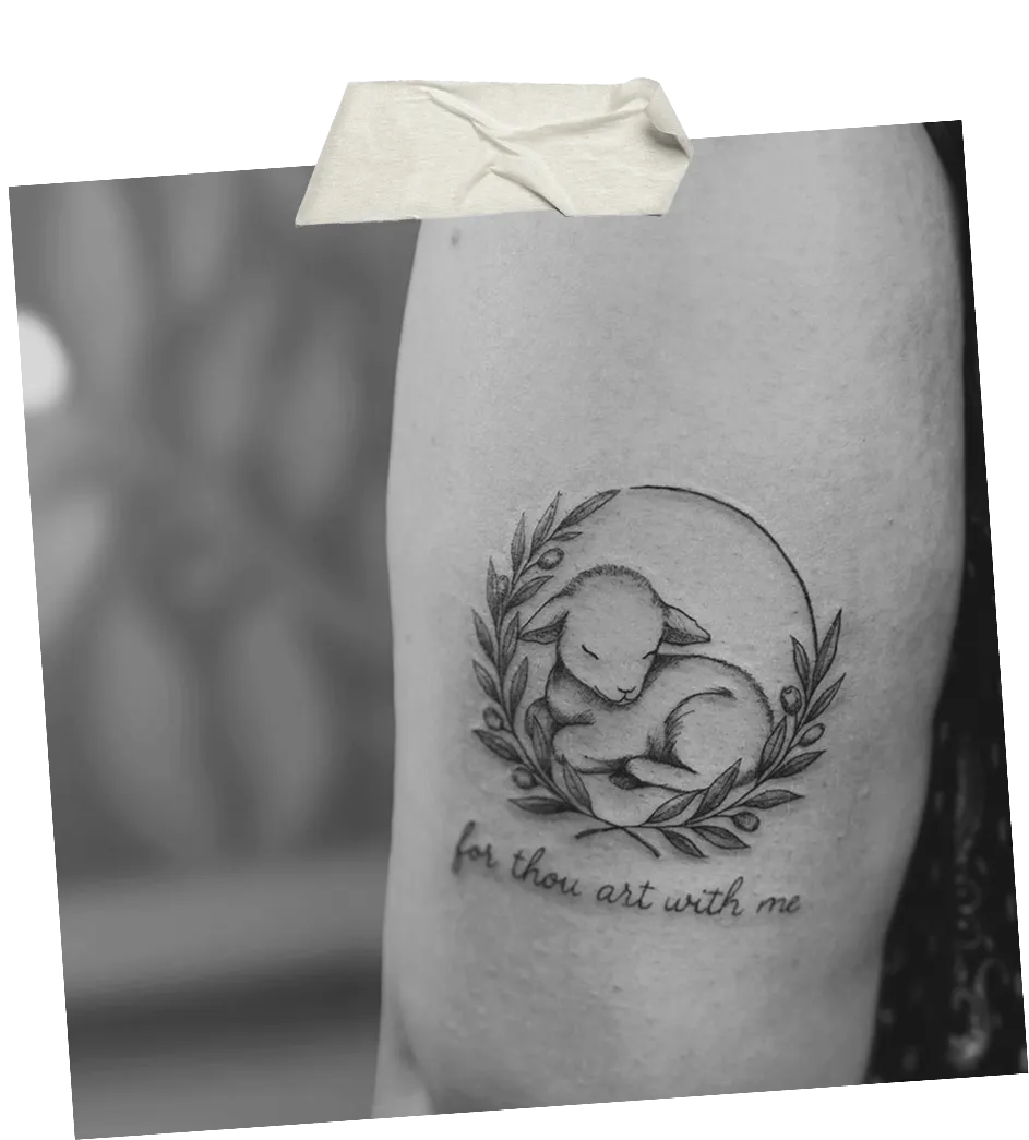 Tattoo of a sleeping lamb surrounded by a wreath of leaves with the text 'for thou art with me' below.