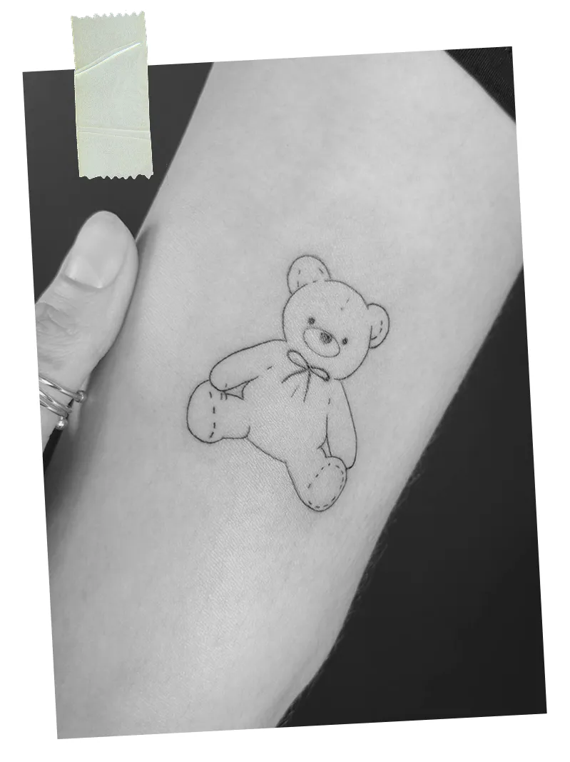 Black and white tattoo of a small teddy bear with a bow on a person's forearm.