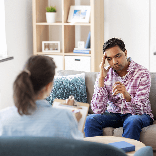 Man in a pink checkered shirt sitting on a couch holding a glass of water and touching his forehead, facing a woman with a clipboard in a therapy session.