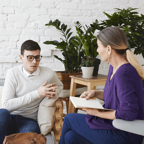 A man with glasses sitting and talking to a woman holding a notebook, with green plants and a white brick wall in the background.