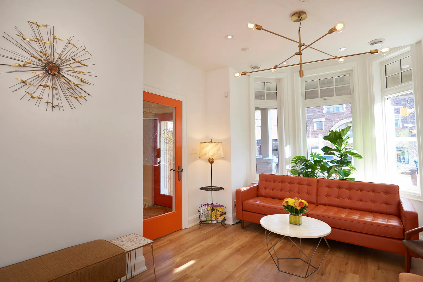 Bright modern office waiting area with orange leather sofa, round marble table with flowers, large windows, and a decorative wall sculpture.