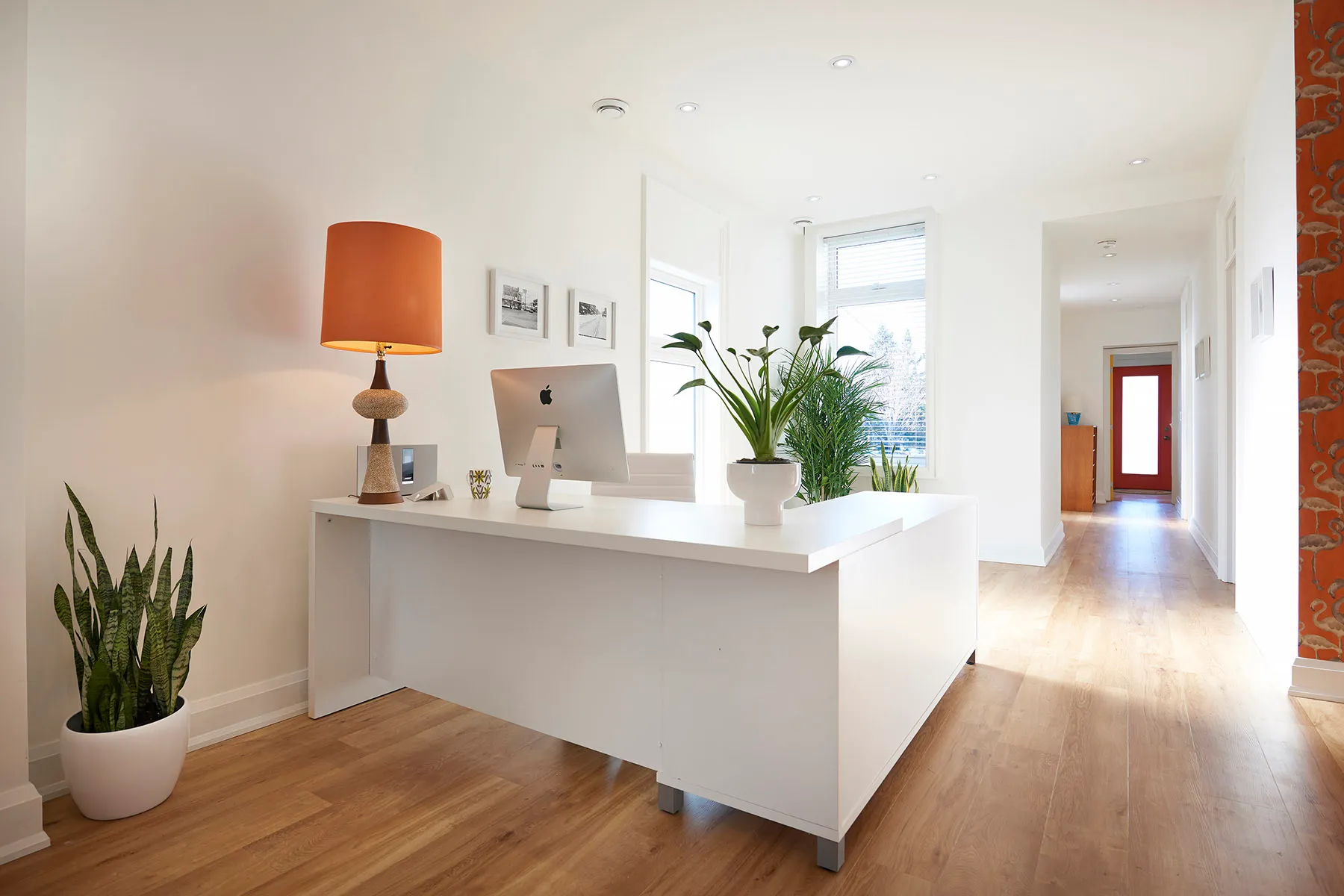 Bright modern office reception area with white desk, computer, orange table lamp, and green plants.