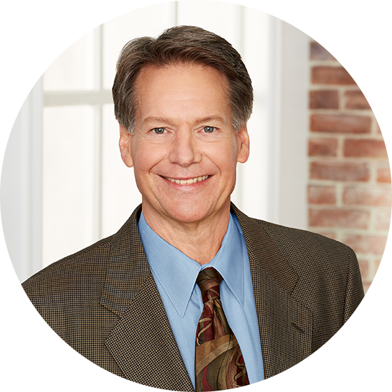 Smiling middle-aged man in a brown checkered blazer, blue shirt, and patterned tie against a bright window and brick wall background.