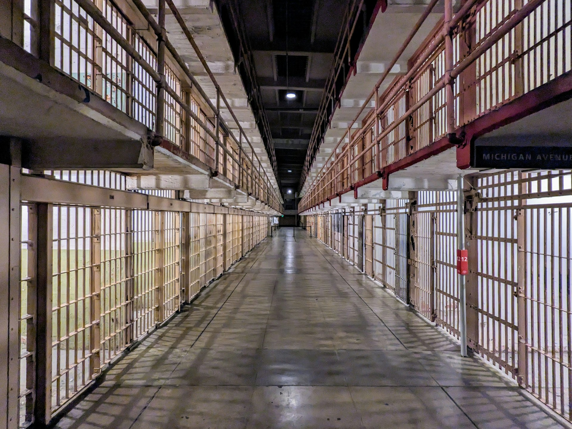 Long prison corridor lined with barred cells on both sides under overhead lighting.
