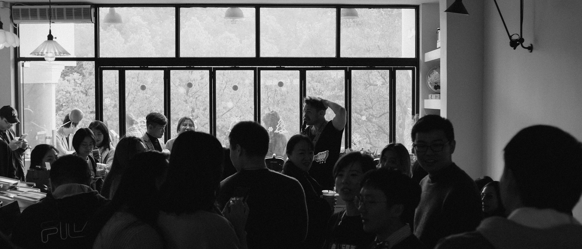Crowded indoor gathering with people socializing in front of large windows.