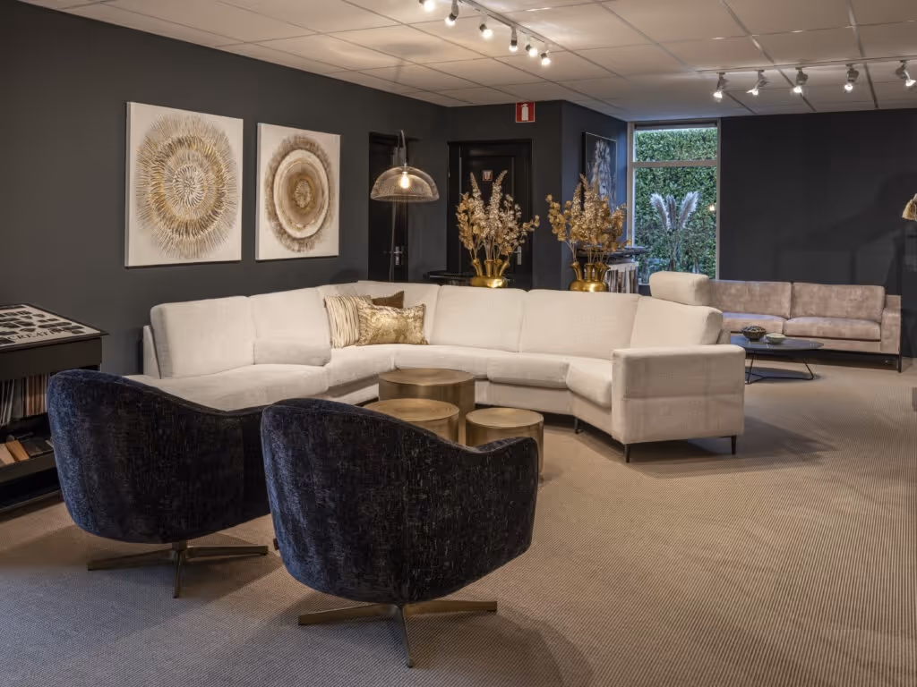 Modern living room with a beige sectional sofa, two dark velvet armchairs, round gold coffee tables, and decorative wall art and plants.