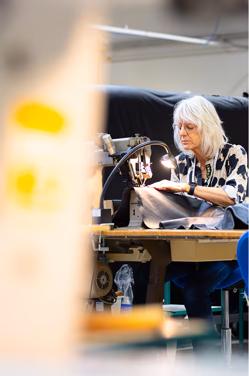Elderly woman with glasses sewing black fabric on an industrial sewing machine in a workshop.