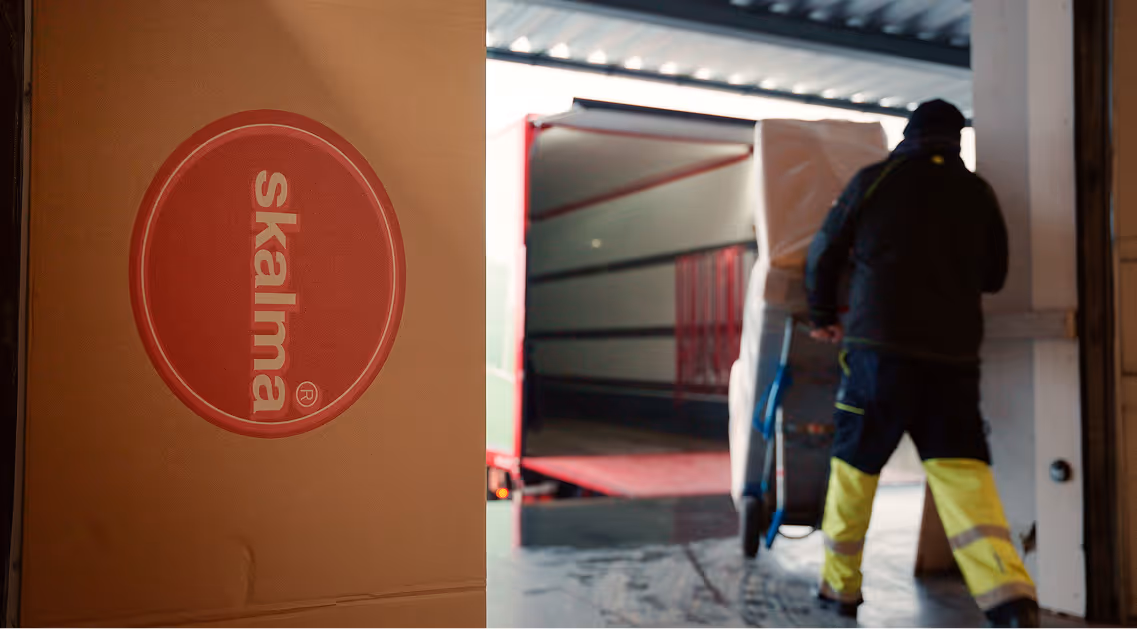 Person in reflective yellow pants moving a large boxed item on a dolly towards a red and white open delivery truck.